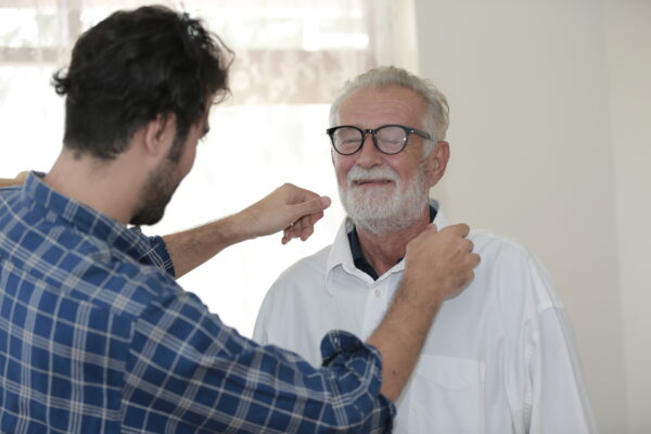 Young caregiver helps older man put on shirt and adjust collar.